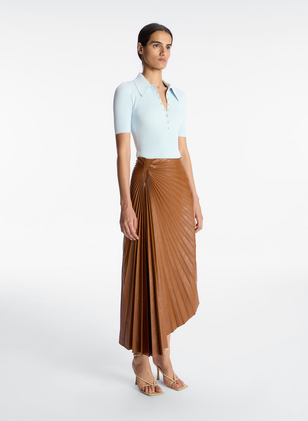 side view of a woman wearing light blue collared shirt with brown pleated vegan leather midi skirt