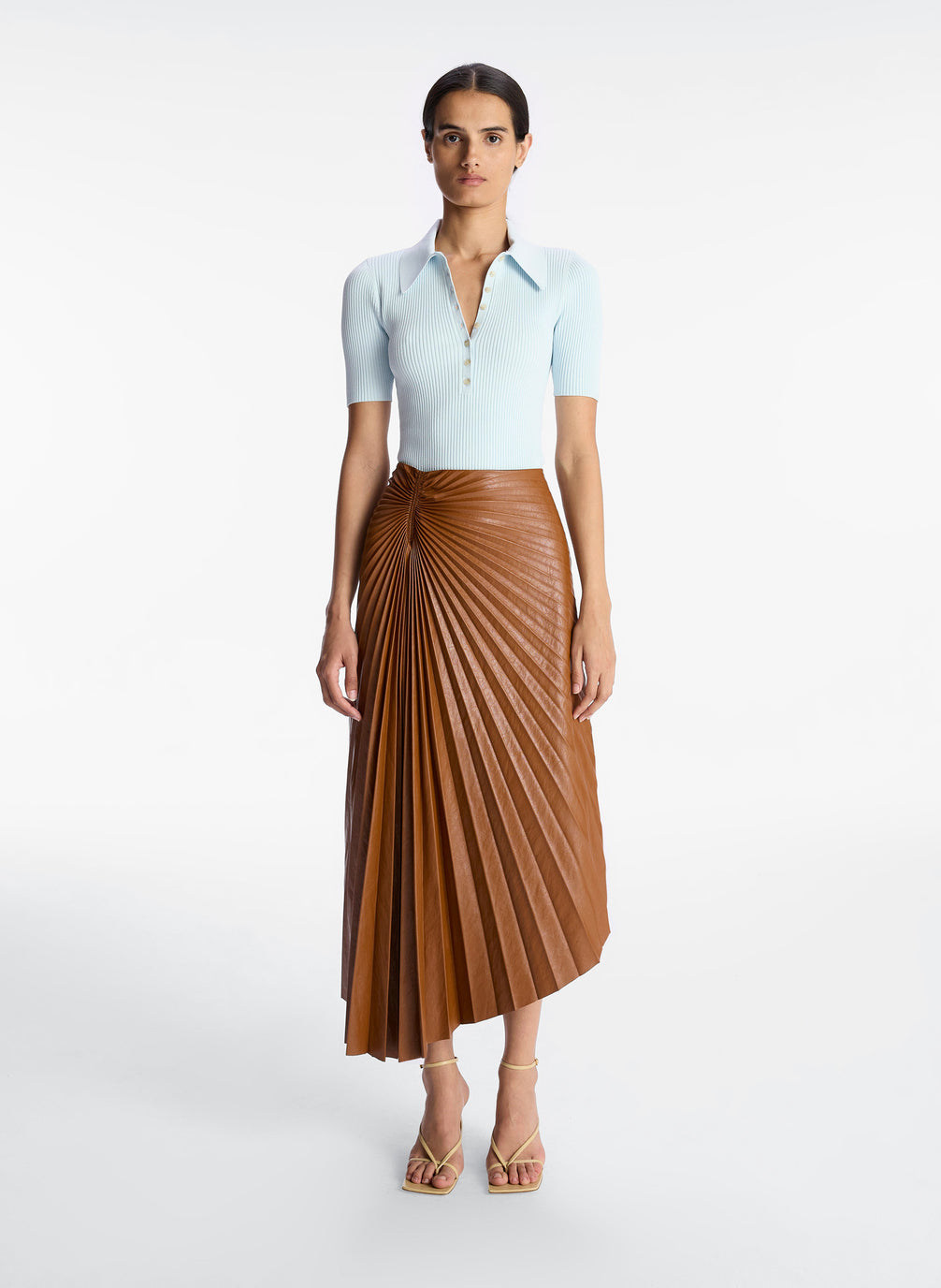 front view of a woman wearing light blue collared shirt with brown pleated vegan leather midi skirt