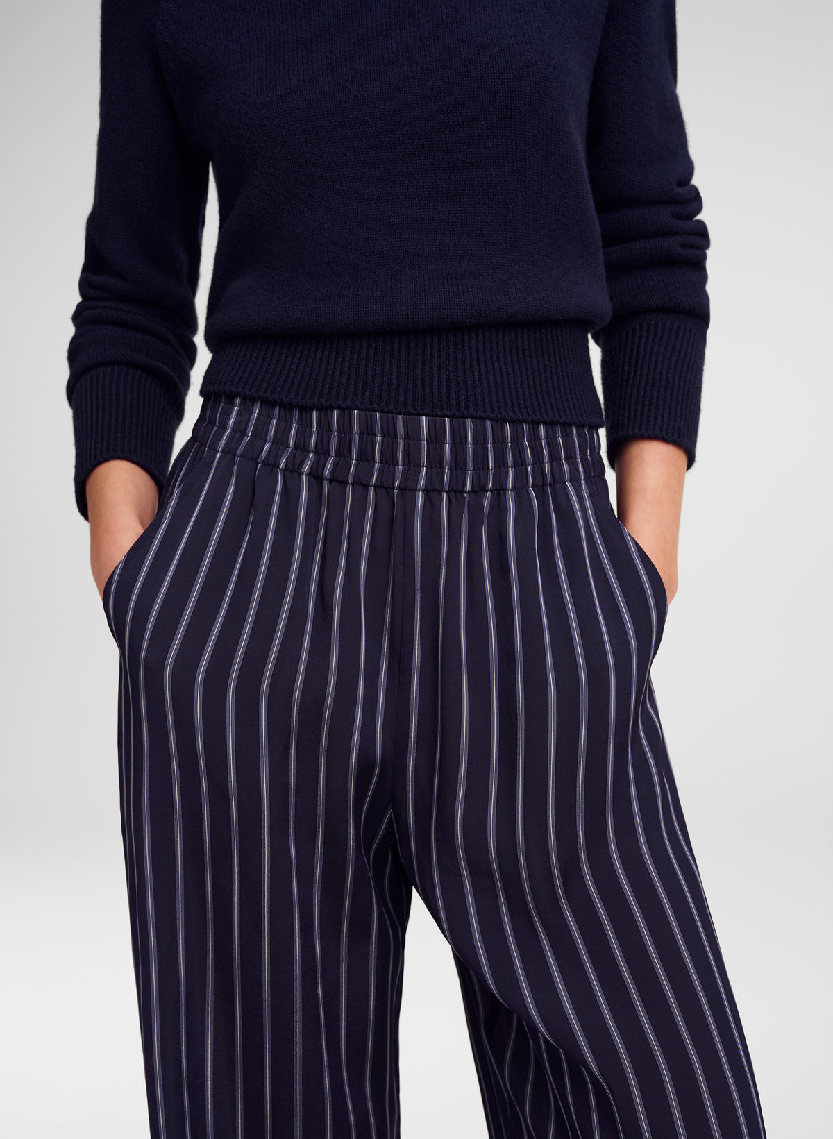 A close-up of a person wearing navy blue pinstriped trousers and a matching navy knit sweater, standing against a light gray studio background.