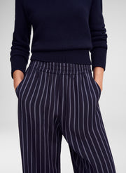 A close-up of a person wearing navy blue pinstriped trousers and a matching navy knit sweater, standing against a light gray studio background.