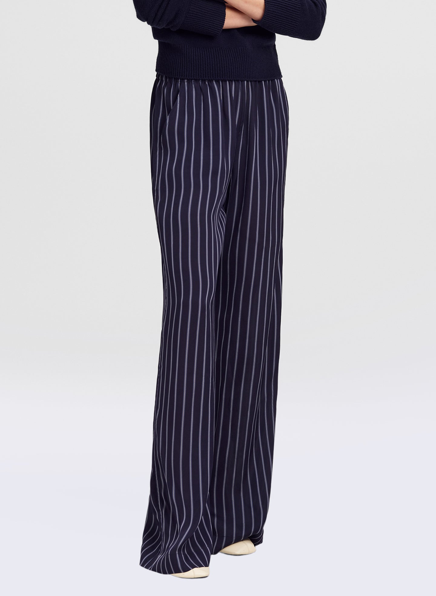 A person wearing navy wide-leg trousers with thin white vertical pinstripes, paired with a dark navy top, shown from the chest down against a light gray studio background.
