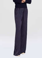 A person wearing navy wide-leg trousers with thin white vertical pinstripes, paired with a dark navy top, shown from the chest down against a light gray studio background.