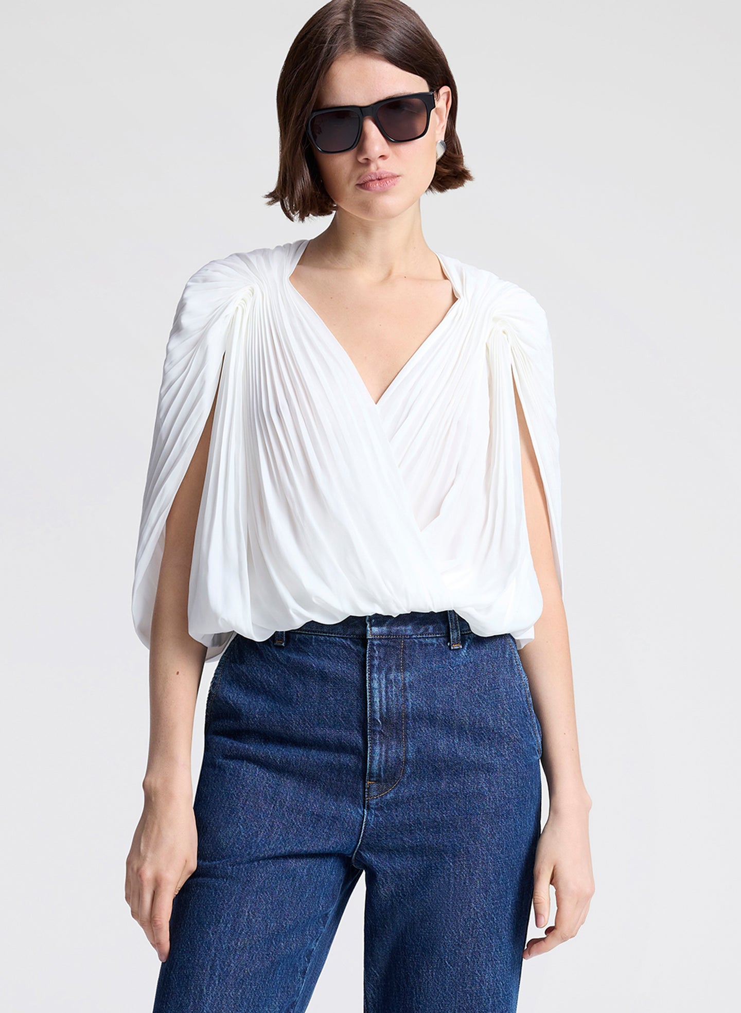 Woman wearing a white top and blue jeans with sunglasses on a light gray background