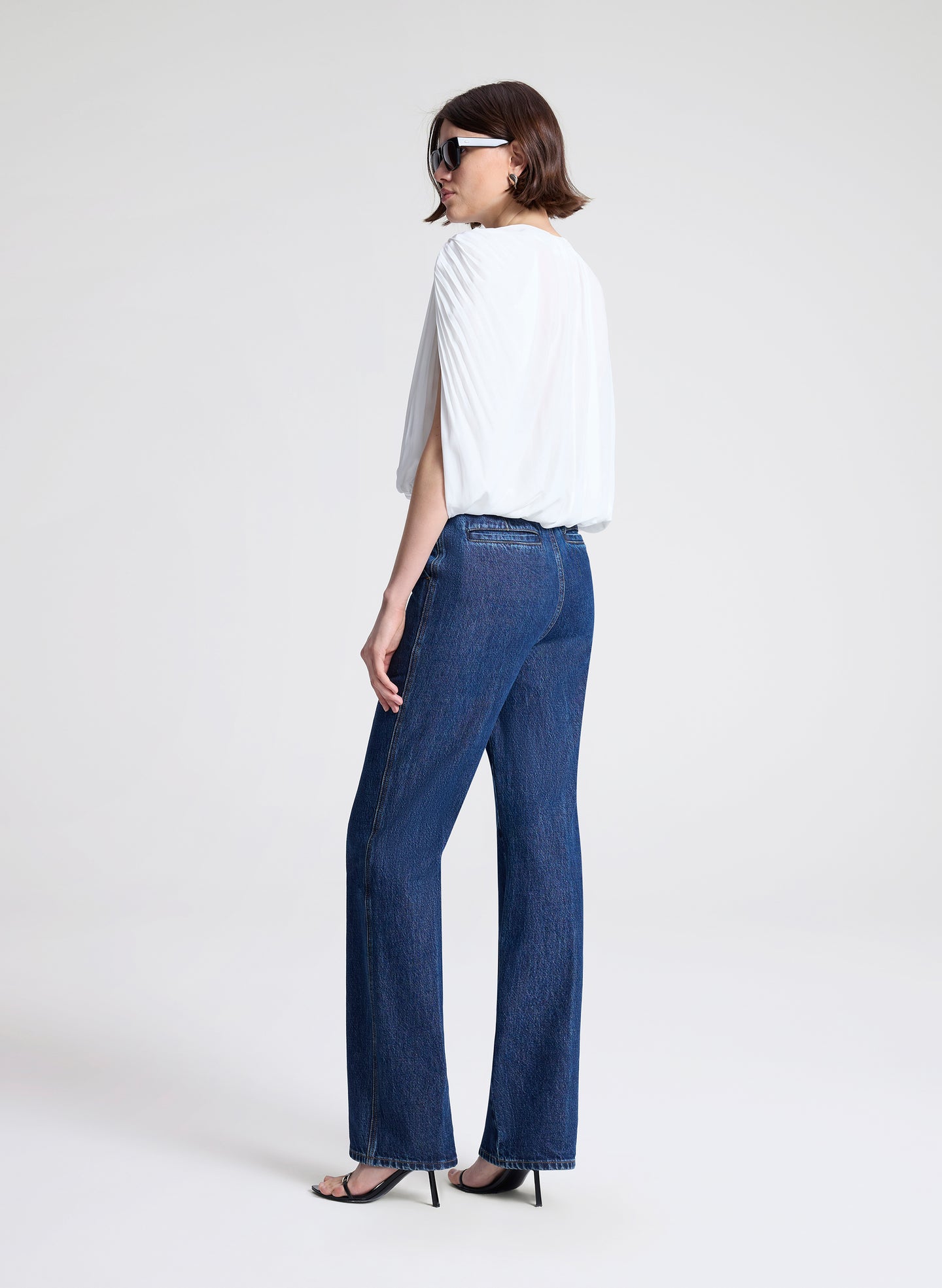 Woman wearing a white top and blue jeans with sunglasses on a light gray background