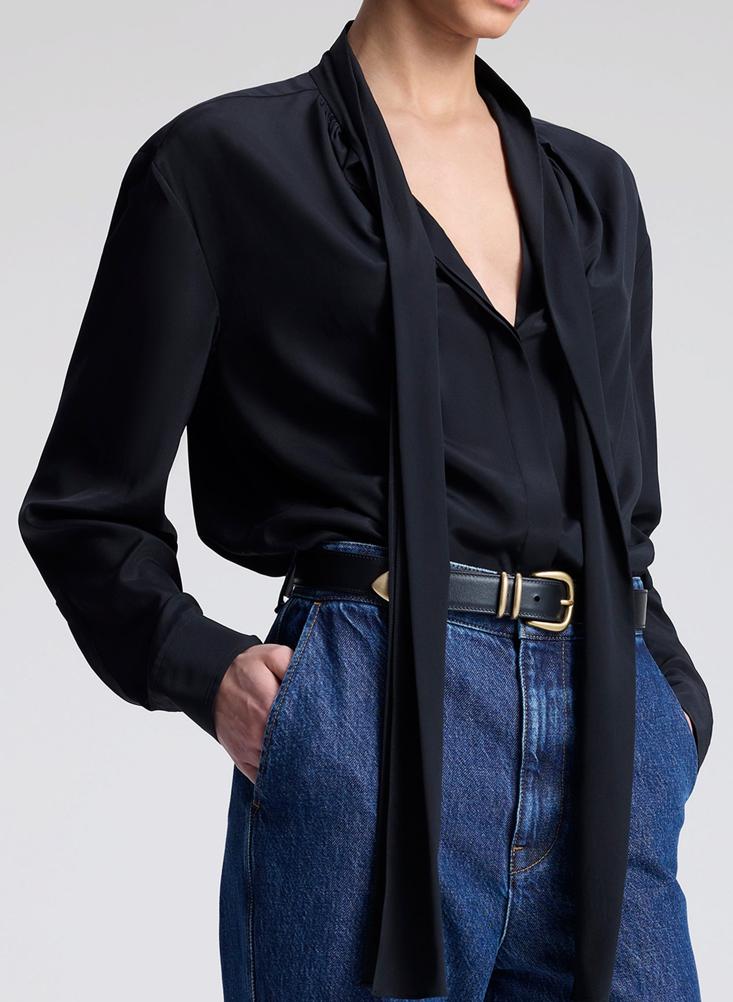 Person wearing a black blouse with a tie detail and blue jeans on a light gray background