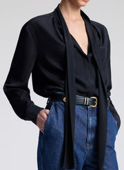 Person wearing a black blouse with a tie detail and blue jeans on a light gray background
