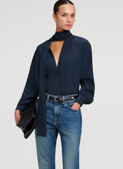 A woman modeling a dark green long-sleeve blouse with a high neck and keyhole cutout, paired with blue jeans and holding a black clutch against a light gray studio background.