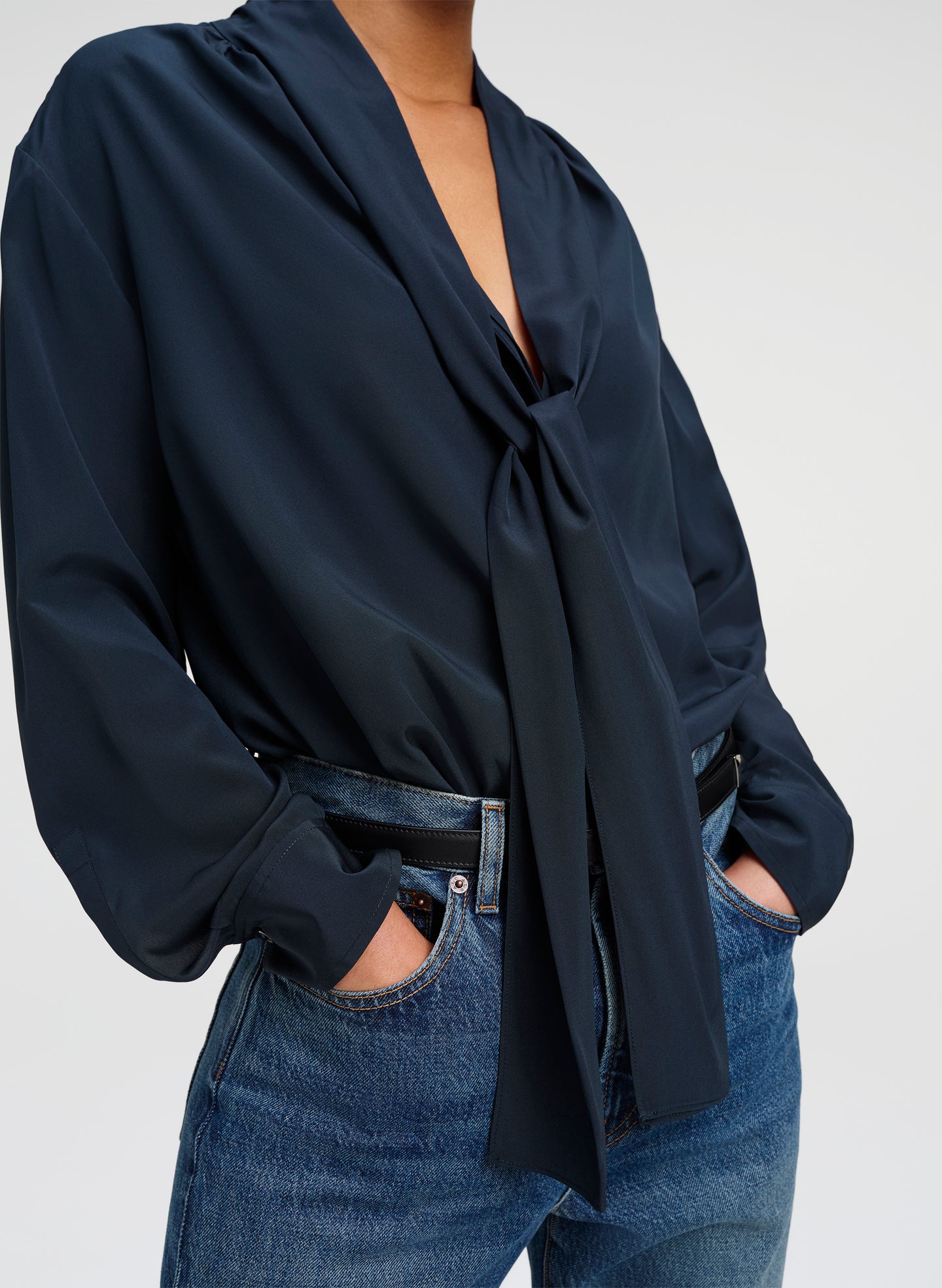 A close-up of a person wearing a dark green blouse with a front tie knot and long sleeves, paired with blue jeans and a black belt, against a light gray studio background.