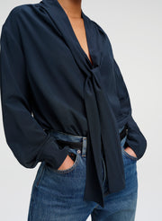 A close-up of a person wearing a dark green blouse with a front tie knot and long sleeves, paired with blue jeans and a black belt, against a light gray studio background.