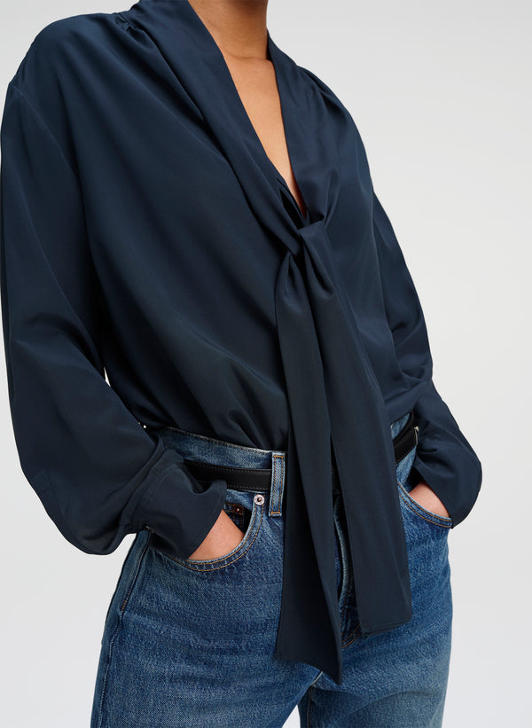 A close-up of a person wearing a dark green blouse with a front tie knot and long sleeves, paired with blue jeans and a black belt, against a light gray studio background.