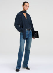 A model stands against a light gray studio background wearing a dark green long-sleeve blouse paired with blue jeans and black shoes, while holding a black clutch.