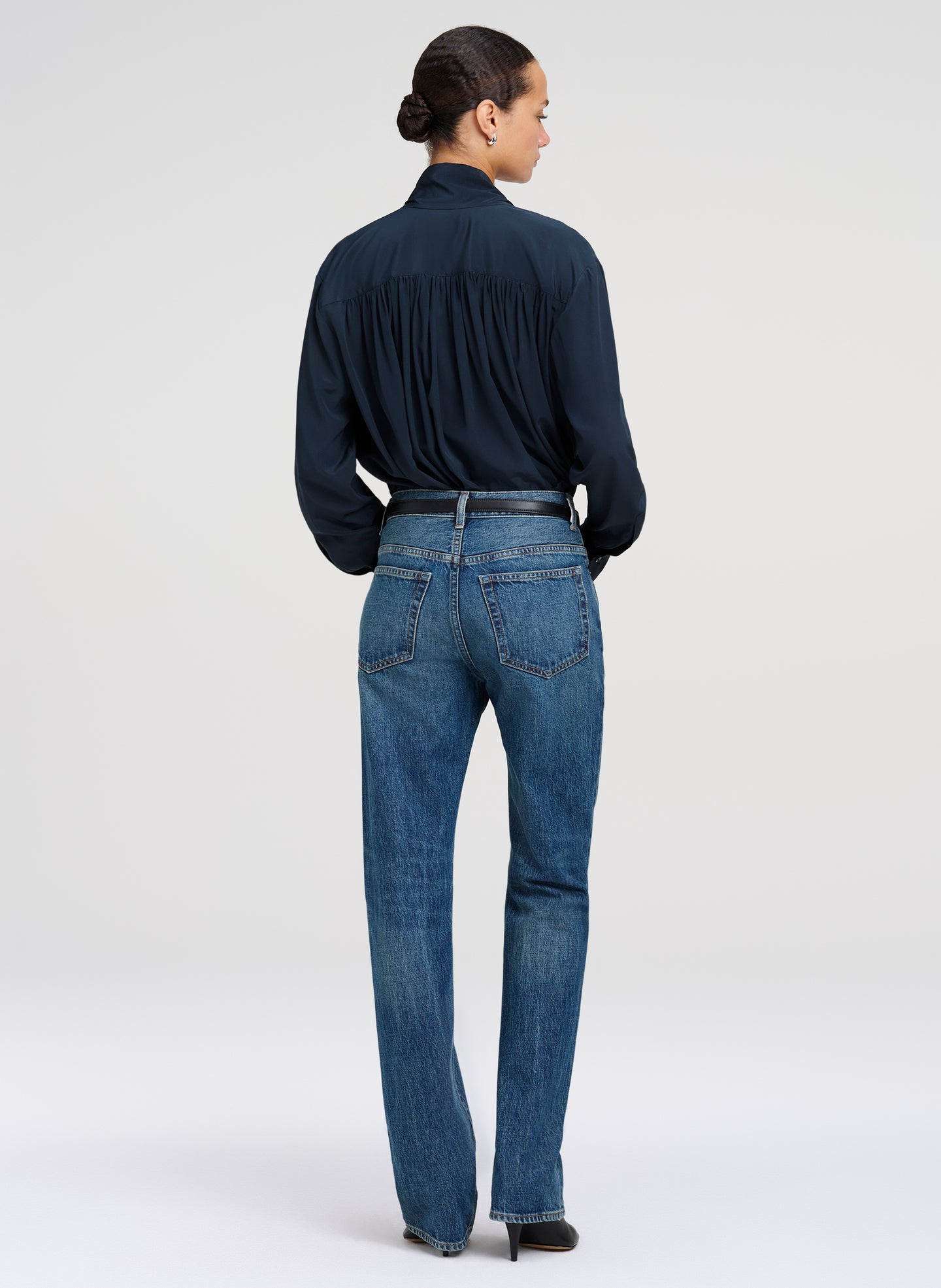 A model shown from the back wearing a dark green long-sleeve blouse tucked into blue denim jeans with a thin black belt, against a light gray studio background.