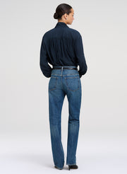 A model shown from the back wearing a dark green long-sleeve blouse tucked into blue denim jeans with a thin black belt, against a light gray studio background.