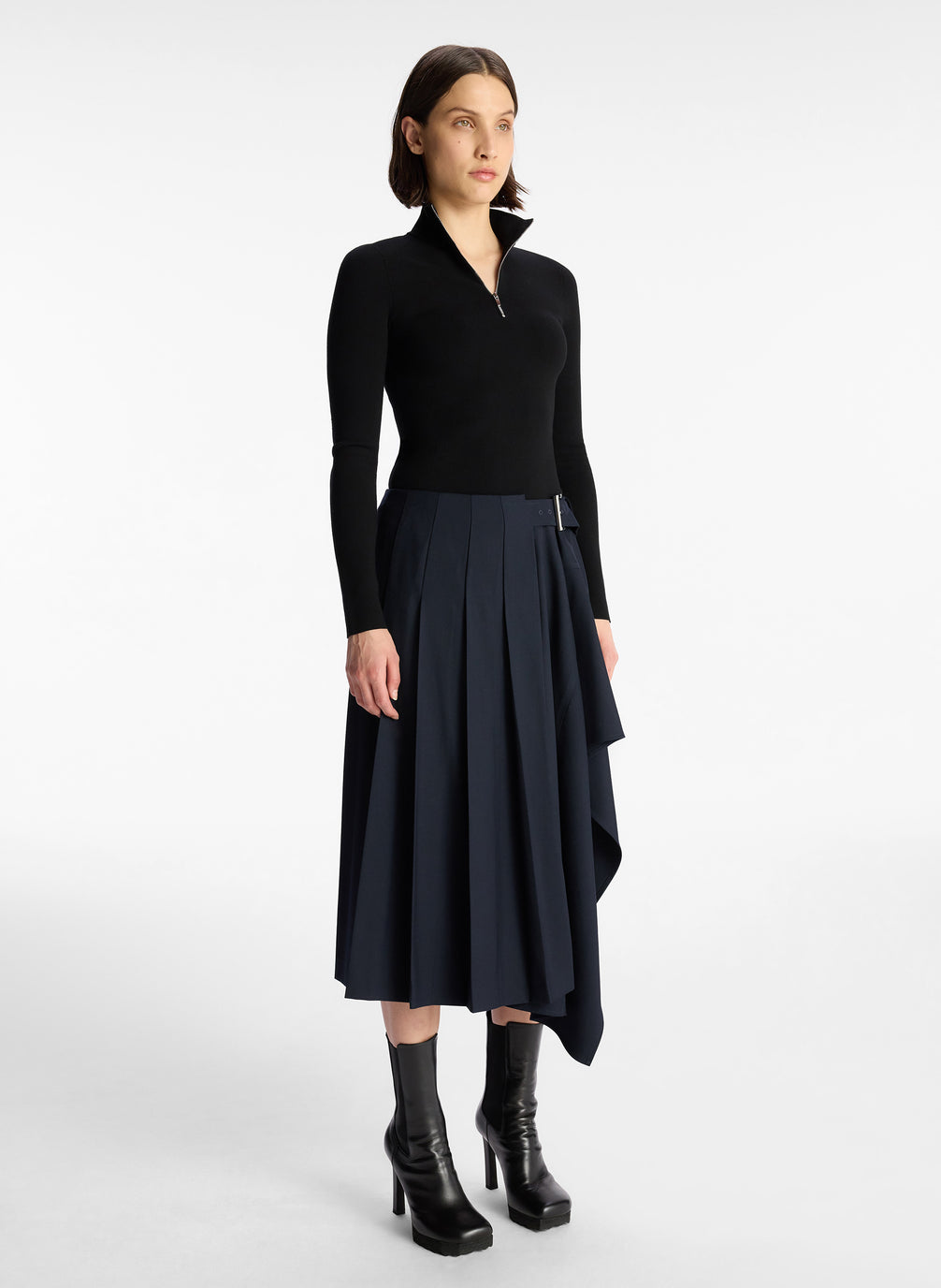 side view of woman in black long sleeve knit top and navy blue pleated skirt
