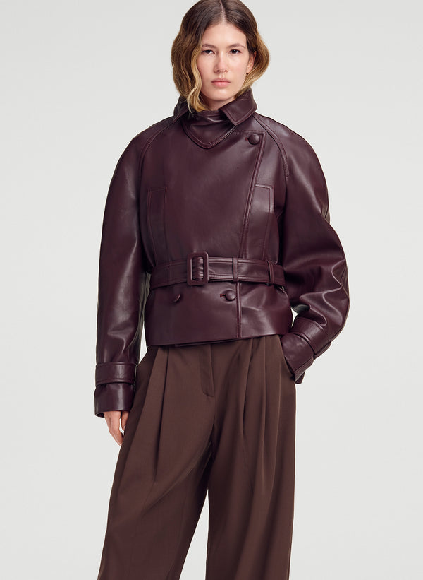 A model wearing a dark burgundy leather cropped jacket with a belted waist and high mock neck, paired with brown wide-leg trousers, against a light gray studio background.