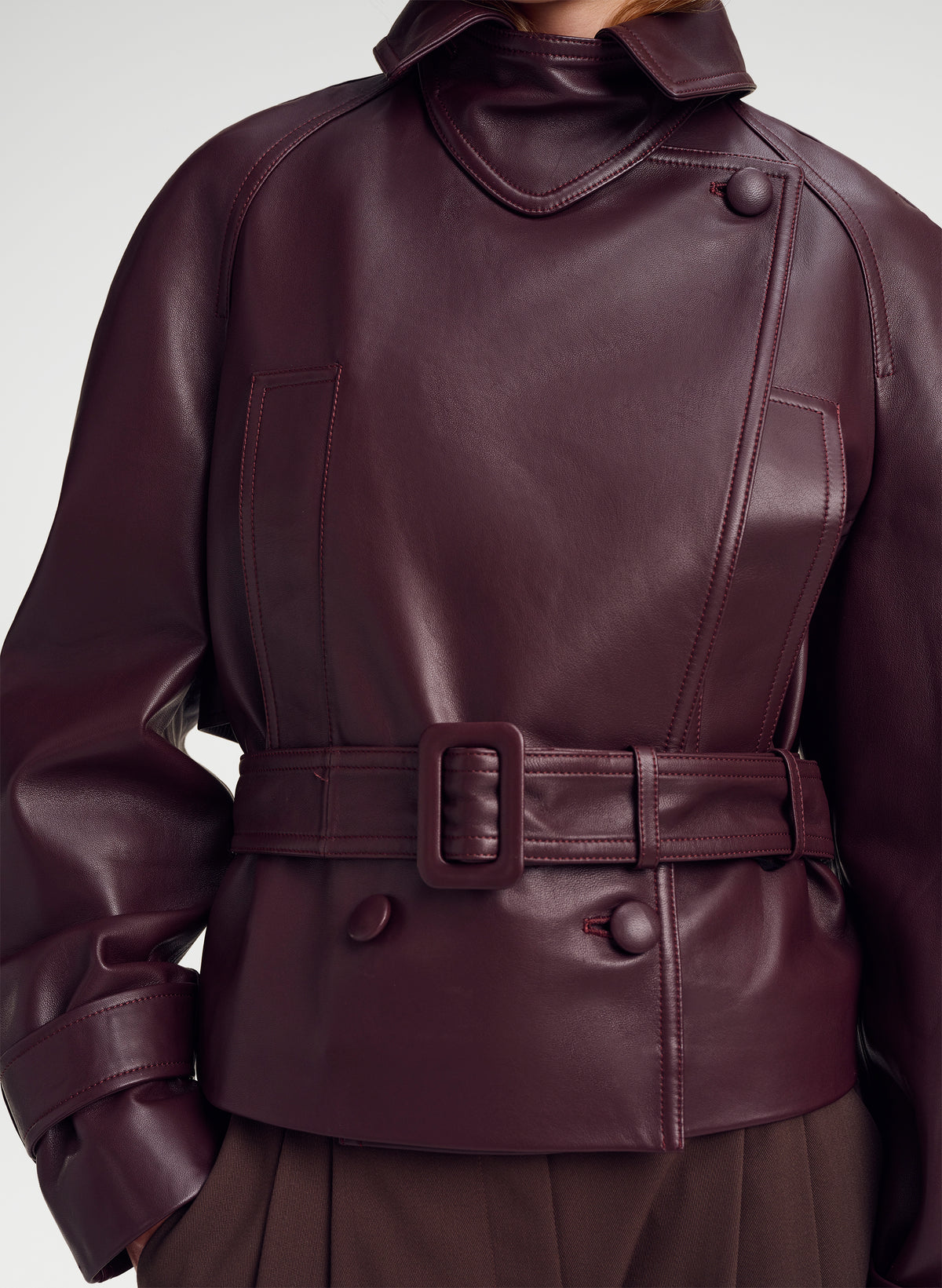 A close-up of a dark burgundy leather jacket with a high collar, belted waist, and structured shoulders, shown on a model against a light gray background.