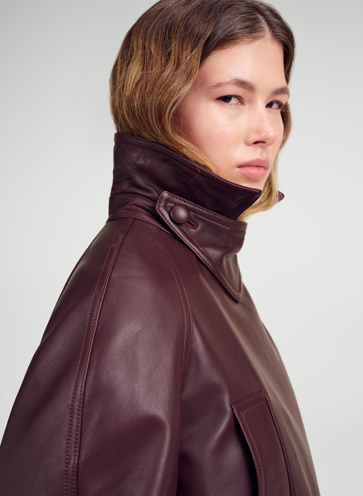 A close-up side profile of a model wearing a dark burgundy leather jacket with a high collar, set against a neutral light gray background.