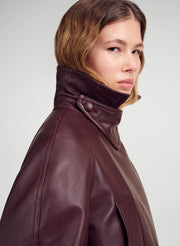 A close-up side profile of a model wearing a dark burgundy leather jacket with a high collar, set against a neutral light gray background.