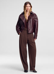 A model stands against a light gray studio background wearing a dark brown leather jacket with contrasting dark red panels, paired with dark brown wide-leg trousers and black pointed shoes.