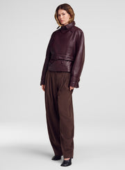 A model stands against a light gray studio background wearing a dark burgundy leather belted jacket and matching dark brown wide-leg trousers.