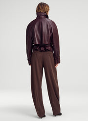 A model shown from the back wearing a dark brown leather jacket with a high collar and ruffled waist detail, paired with matching wide-leg trousers, against a light gray studio background.