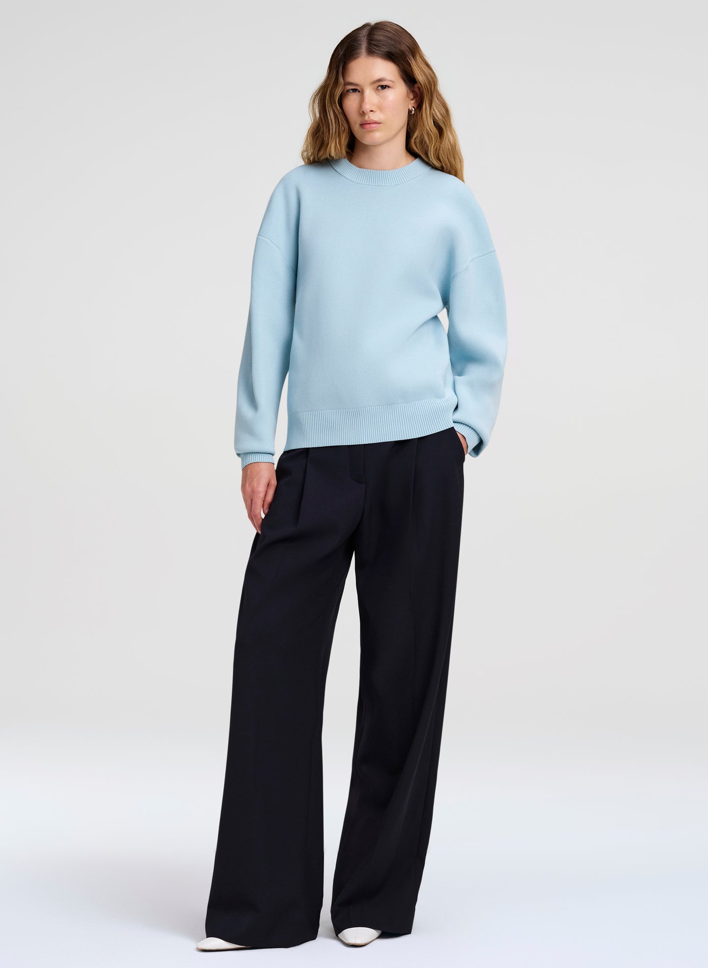 A woman modeling a light blue crewneck sweater and dark blue wide-leg trousers against a plain light gray studio background.