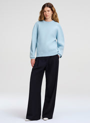 A woman modeling a light blue crewneck sweater and dark blue wide-leg trousers against a plain light gray studio background.