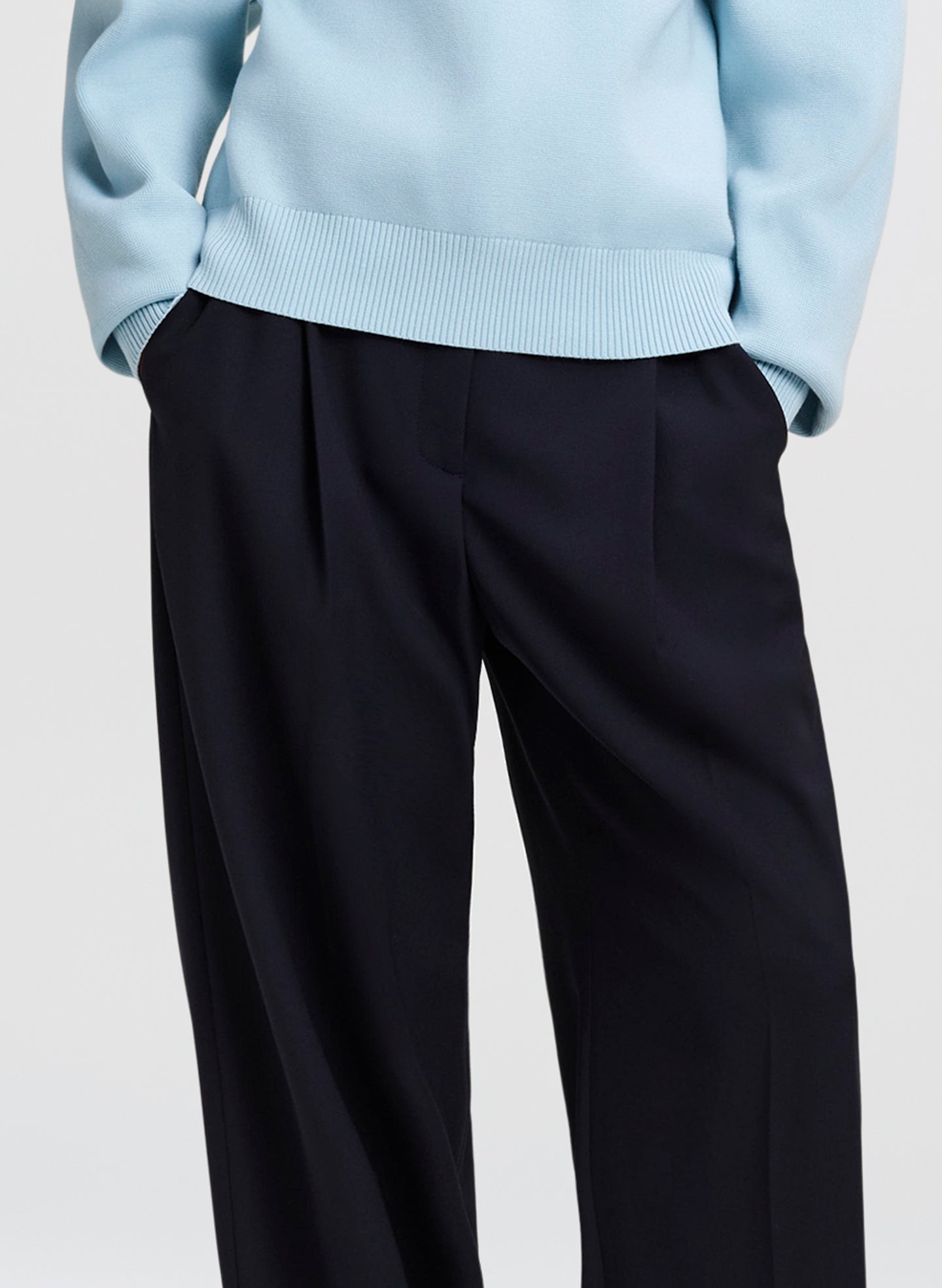 Close-up of a person wearing dark navy wide-leg trousers and a light blue ribbed sweater, cropped from the chest down against a plain light gray background.