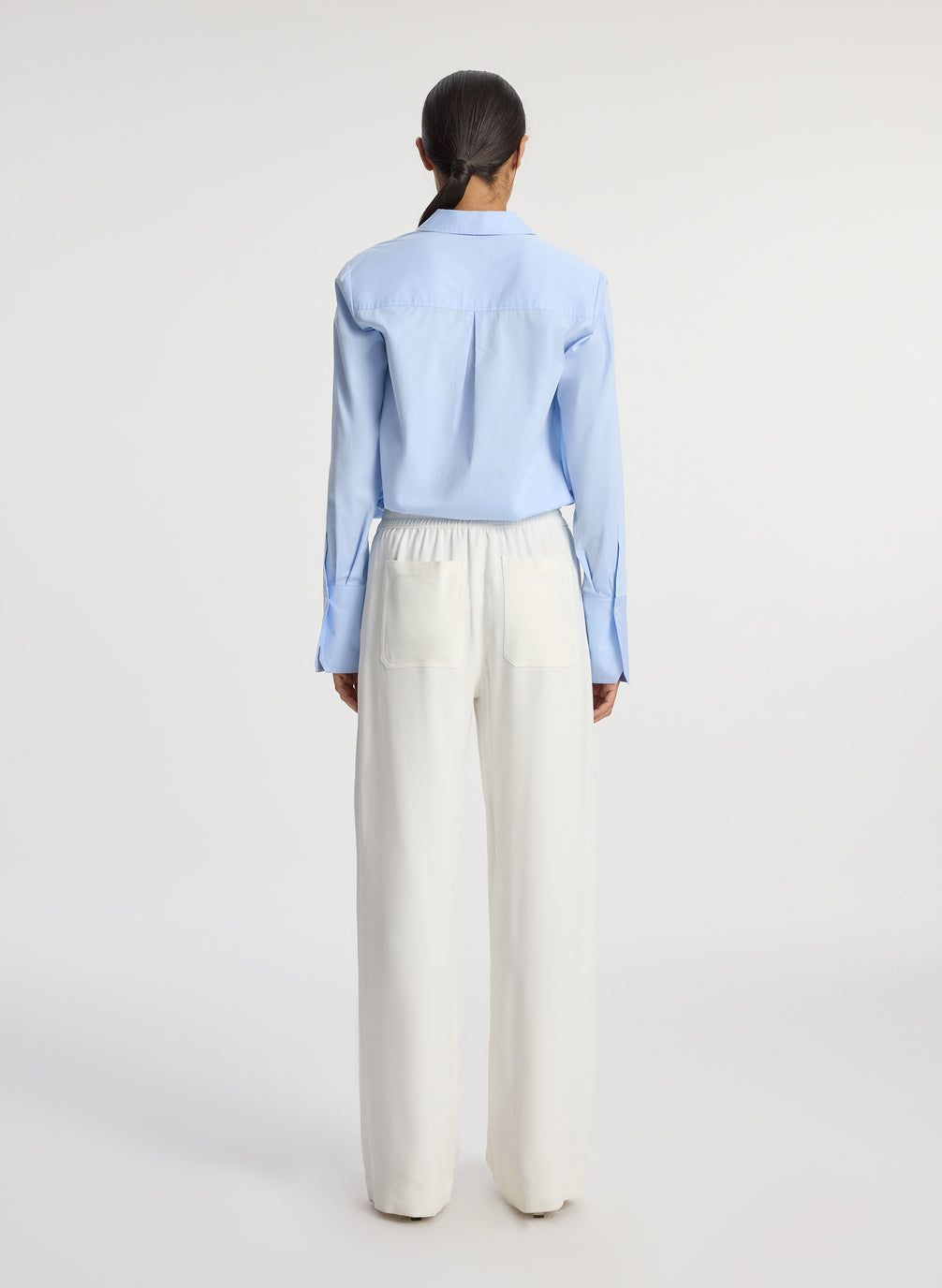 back view of woman wearing light blue button down collared shirt and white pants