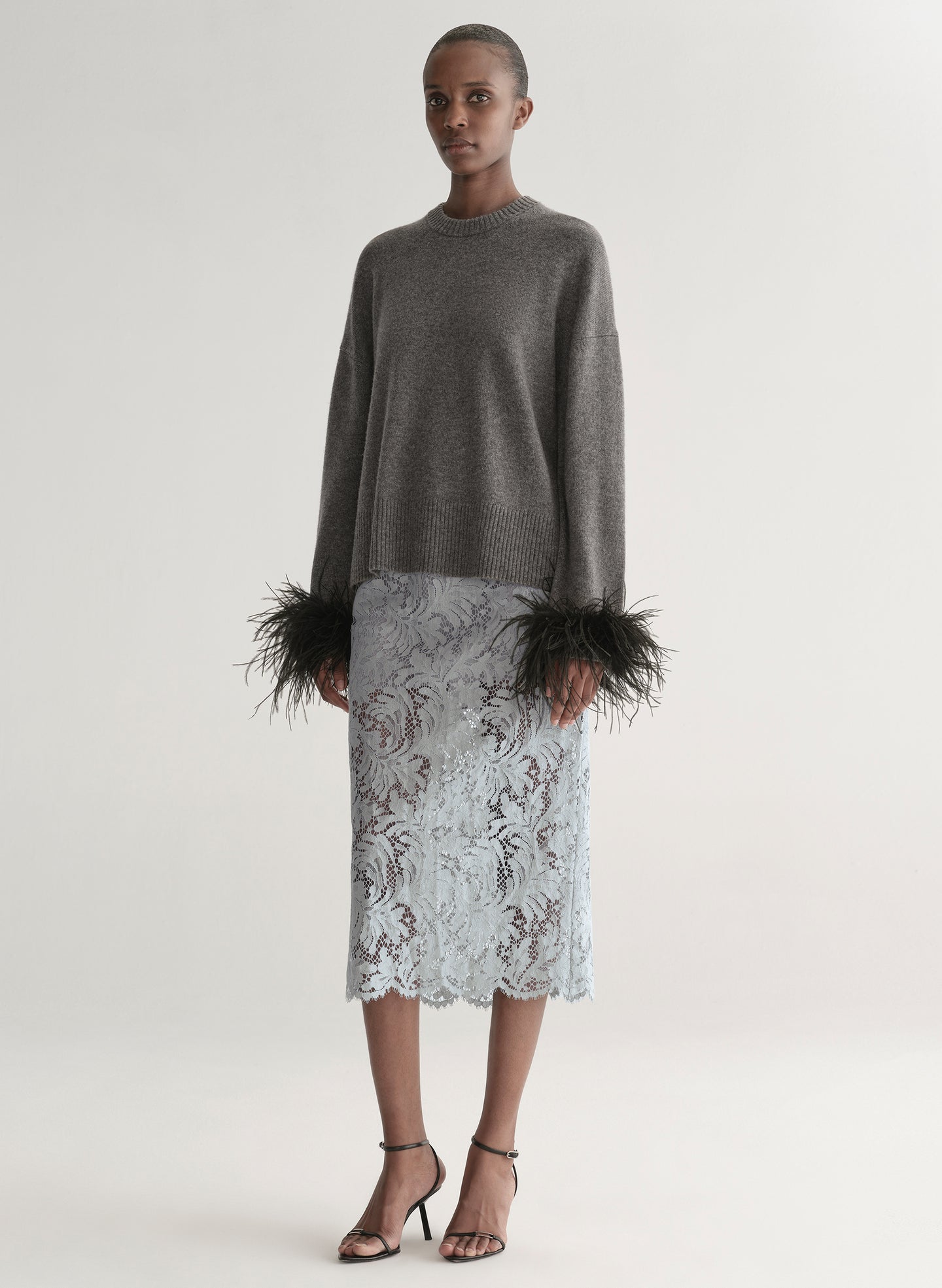 Woman wearing a grey sweater with feather details  and a lace skirt on a plain background