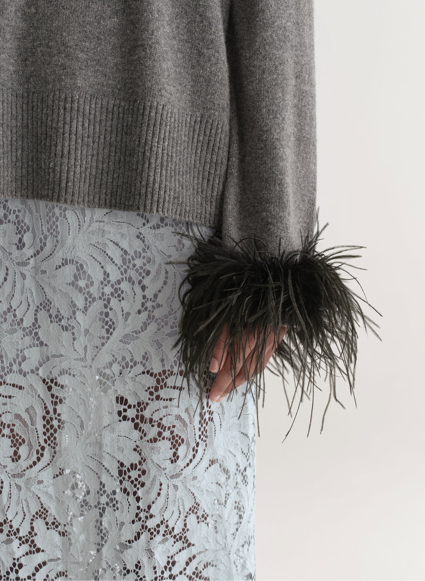 Woman wearing a grey sweater with feather details  and a lace skirt on a plain background