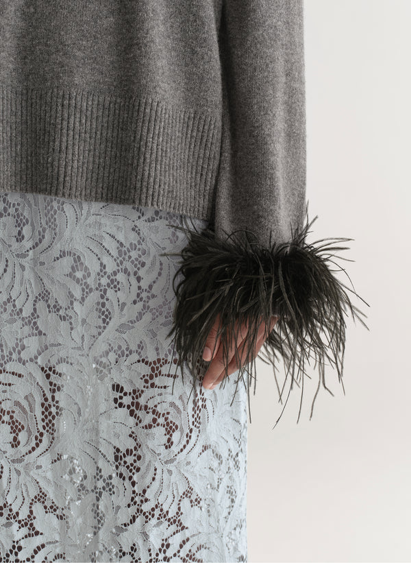 Woman wearing a grey sweater with feather details  and a lace skirt on a plain background