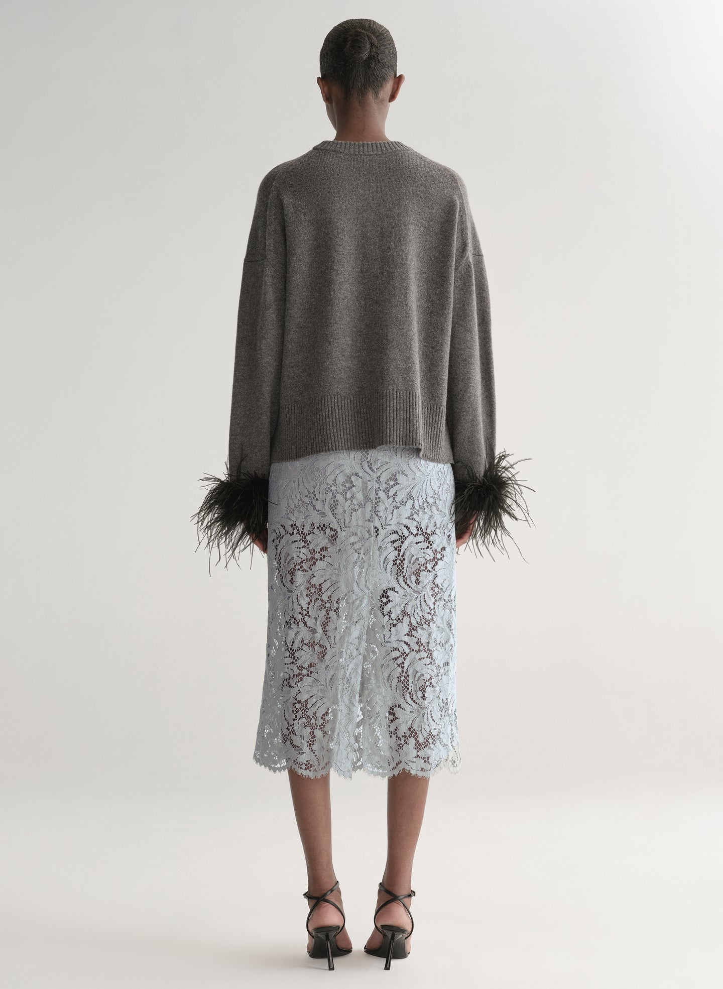Woman wearing a grey sweater with feather details  and a lace skirt on a plain background