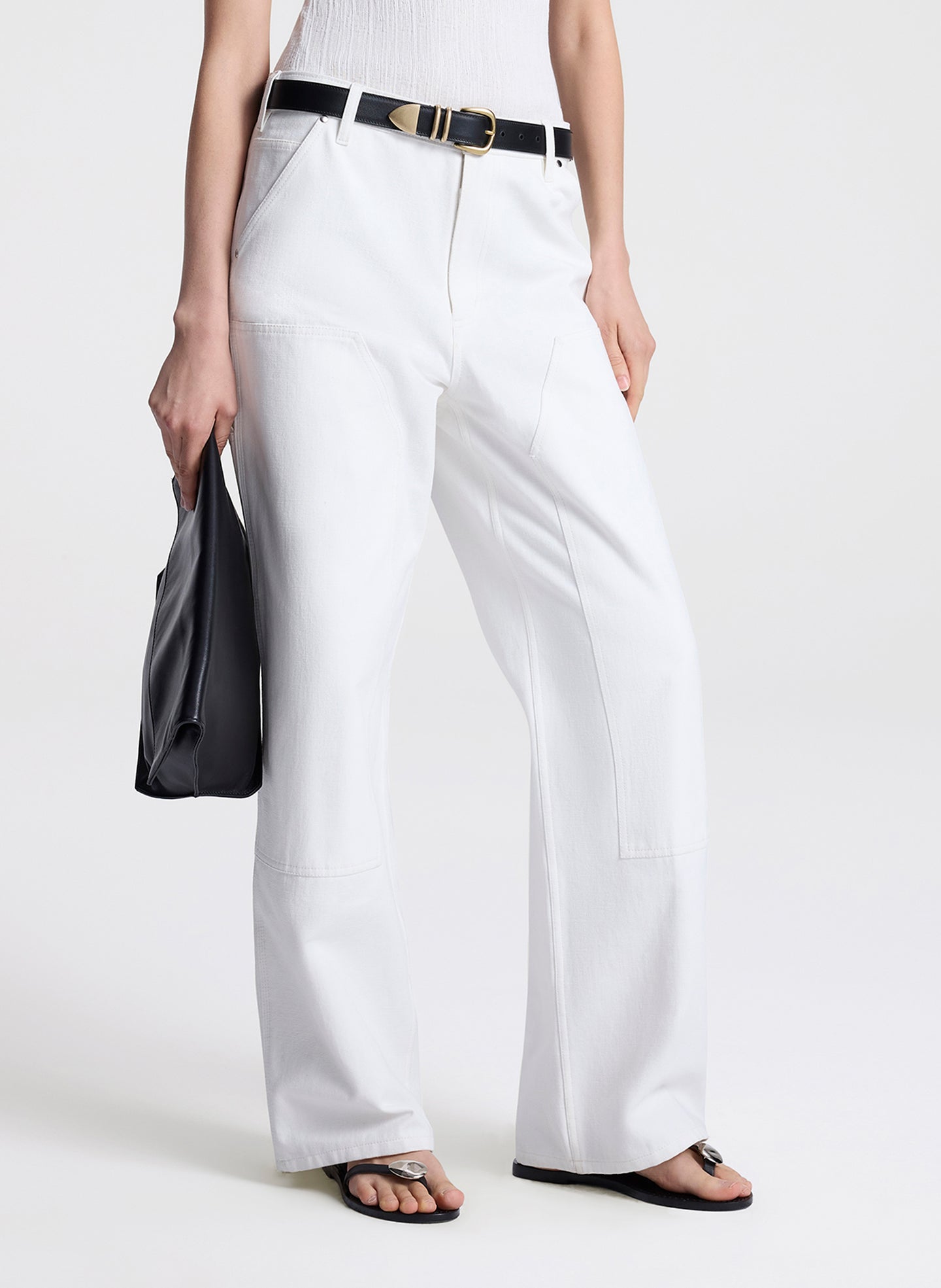 Person wearing white pants with a belt and holding a black clutch against a white background