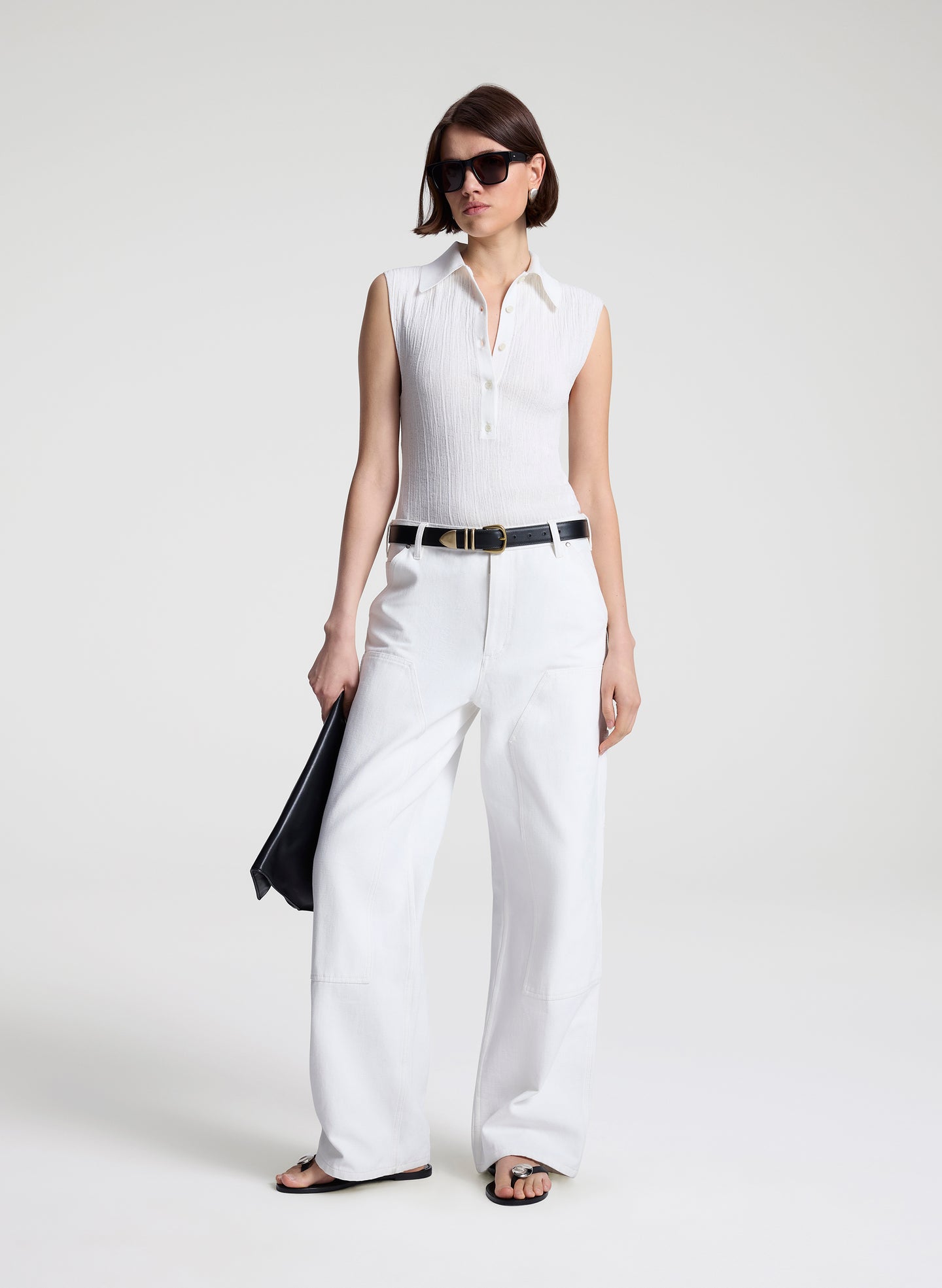 Woman wearing a white sleeveless shirt and white pants with a black belt on a white background