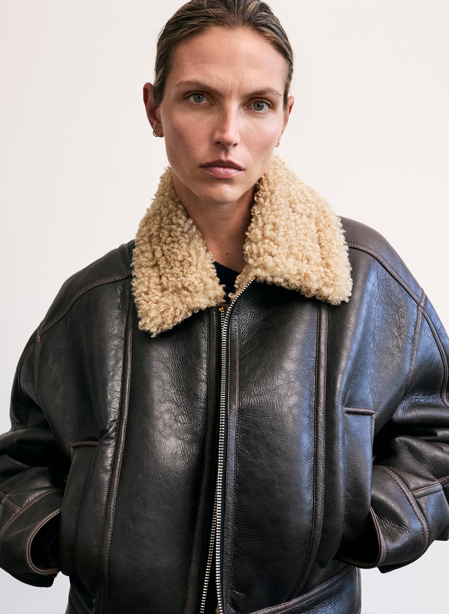 Person wearing a black faux leather jacket with beige shearling collar on a plain background